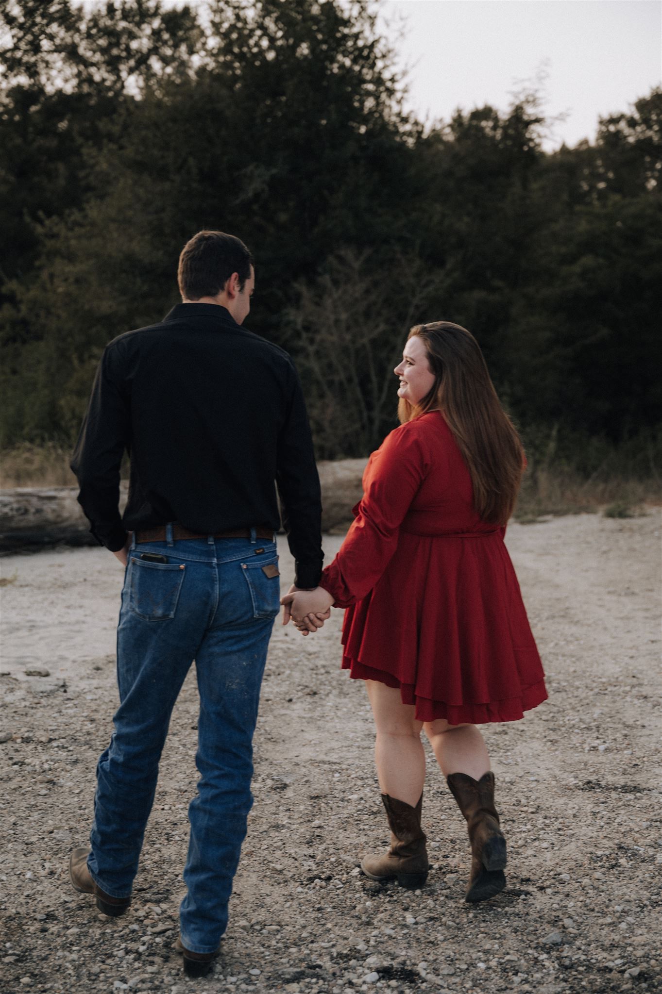 Two people hold hands while walking on a sandy path surrounded by trees.
