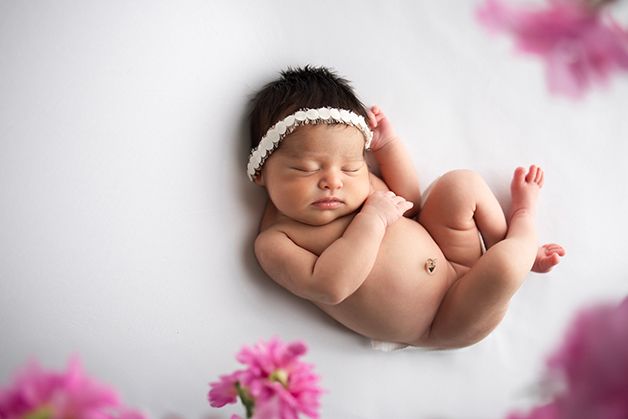 Newborn baby girl curled on a white blanket with pink flowers around the frame.