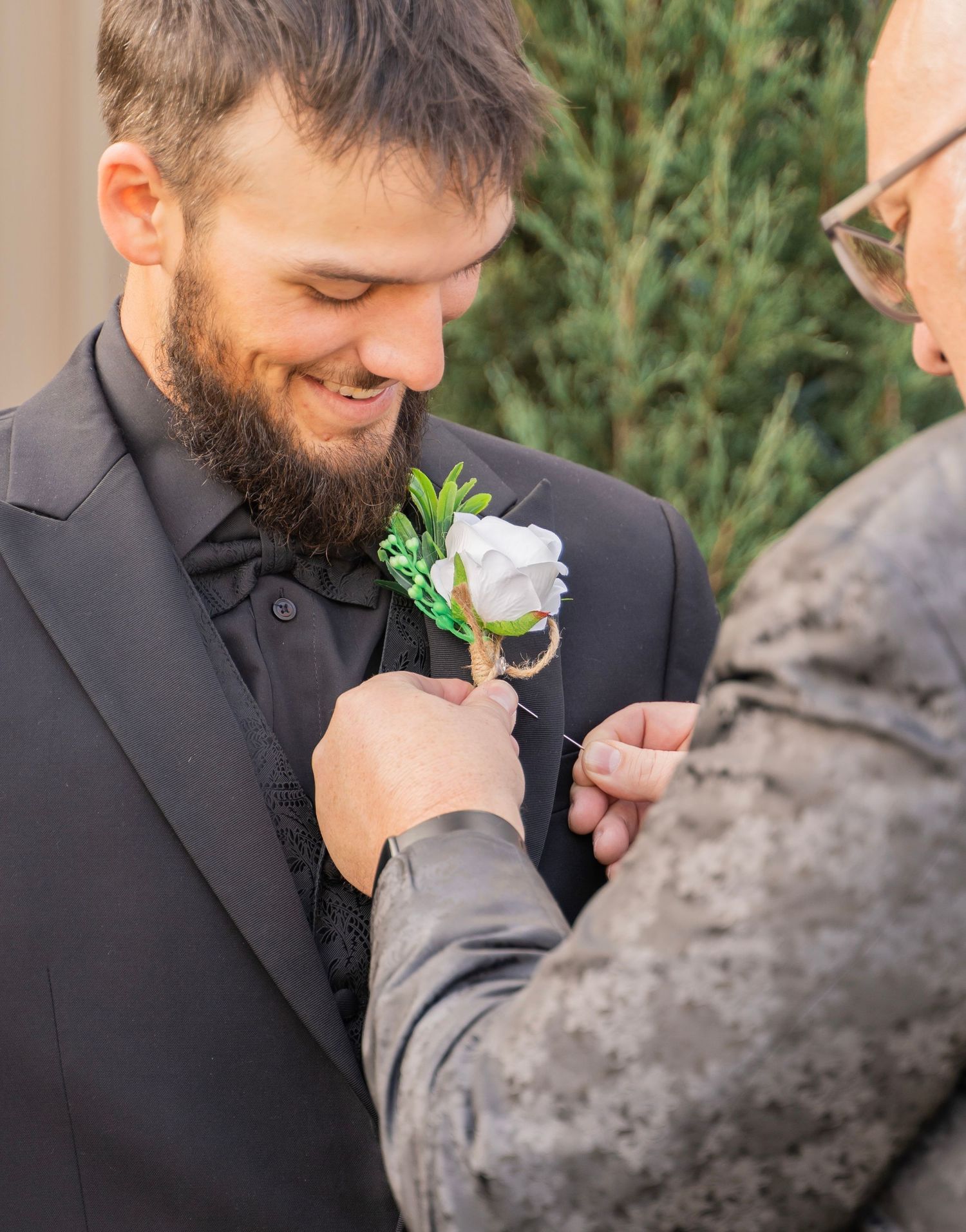 Smiling bearded groom having a white rose boutonniere pinned to his black suit jacket.