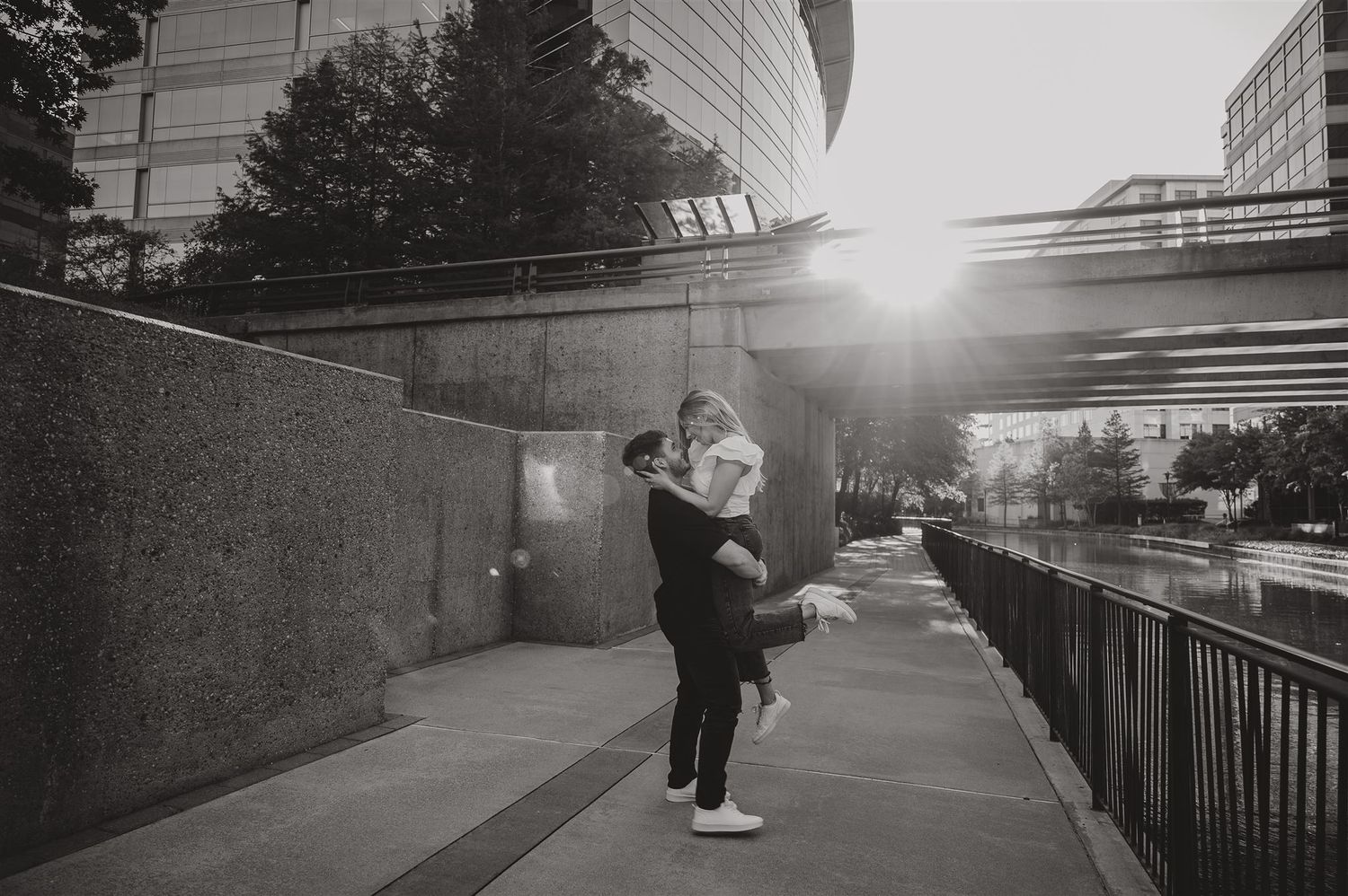 A romantic couple embrace on an urban walkway at sunset near modern buildings.