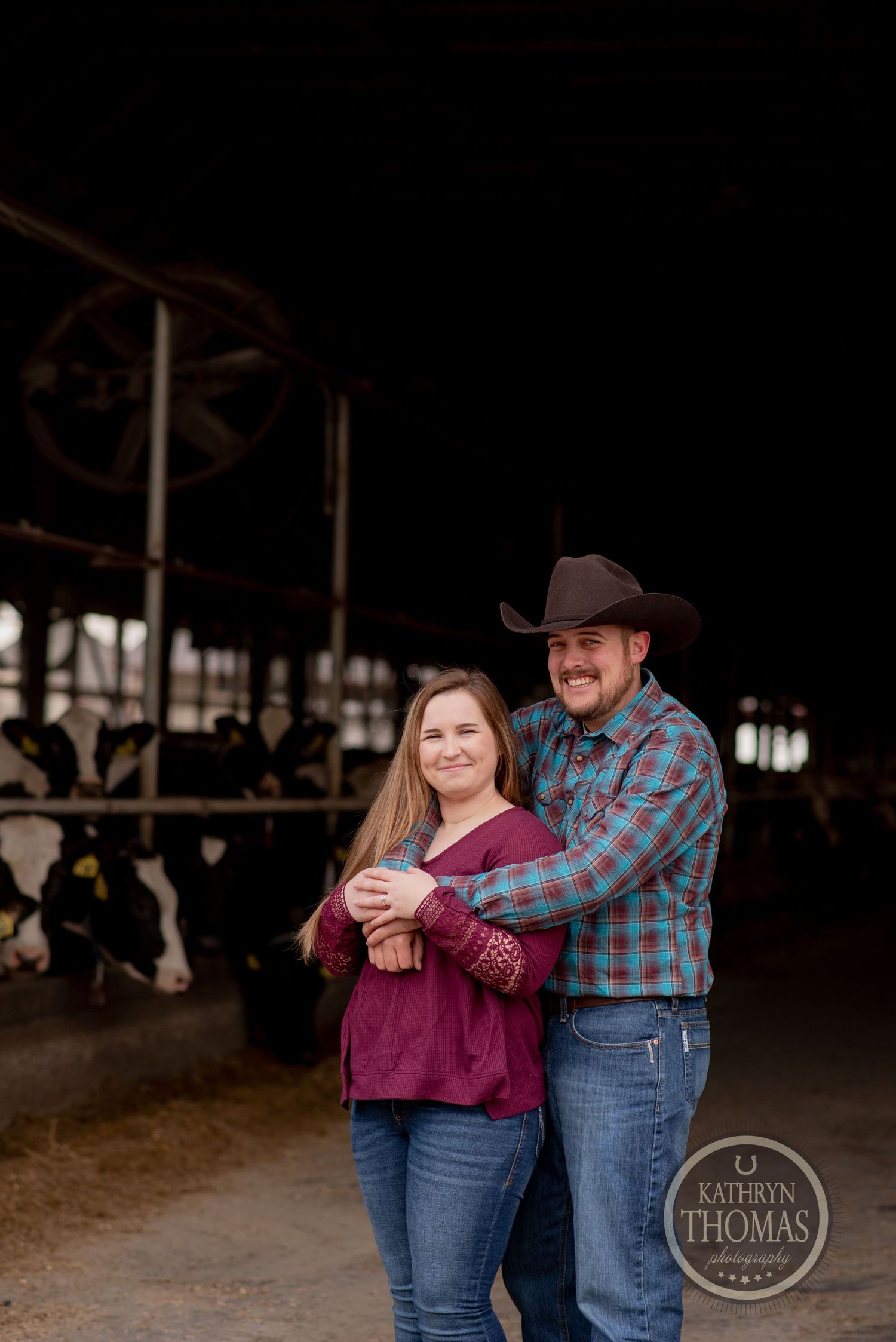 Couple smiling and embracing in dairy barn while wearing country western clothing.