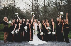 Wedding party in black dresses holding white bouquets poses with bride in elegant white gown outdoors.