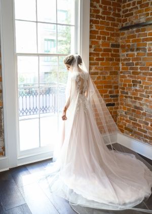 Bride in flowing lace gown and veil stands by bright window in rustic brick room.