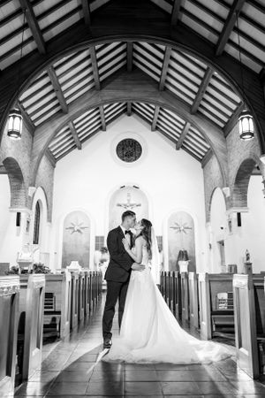 Black and white wedding portrait in church aisle with soaring wooden beams and arched ceiling.