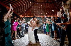 Wedding couple kisses during sparkler exit while guests cheer in rustic venue.