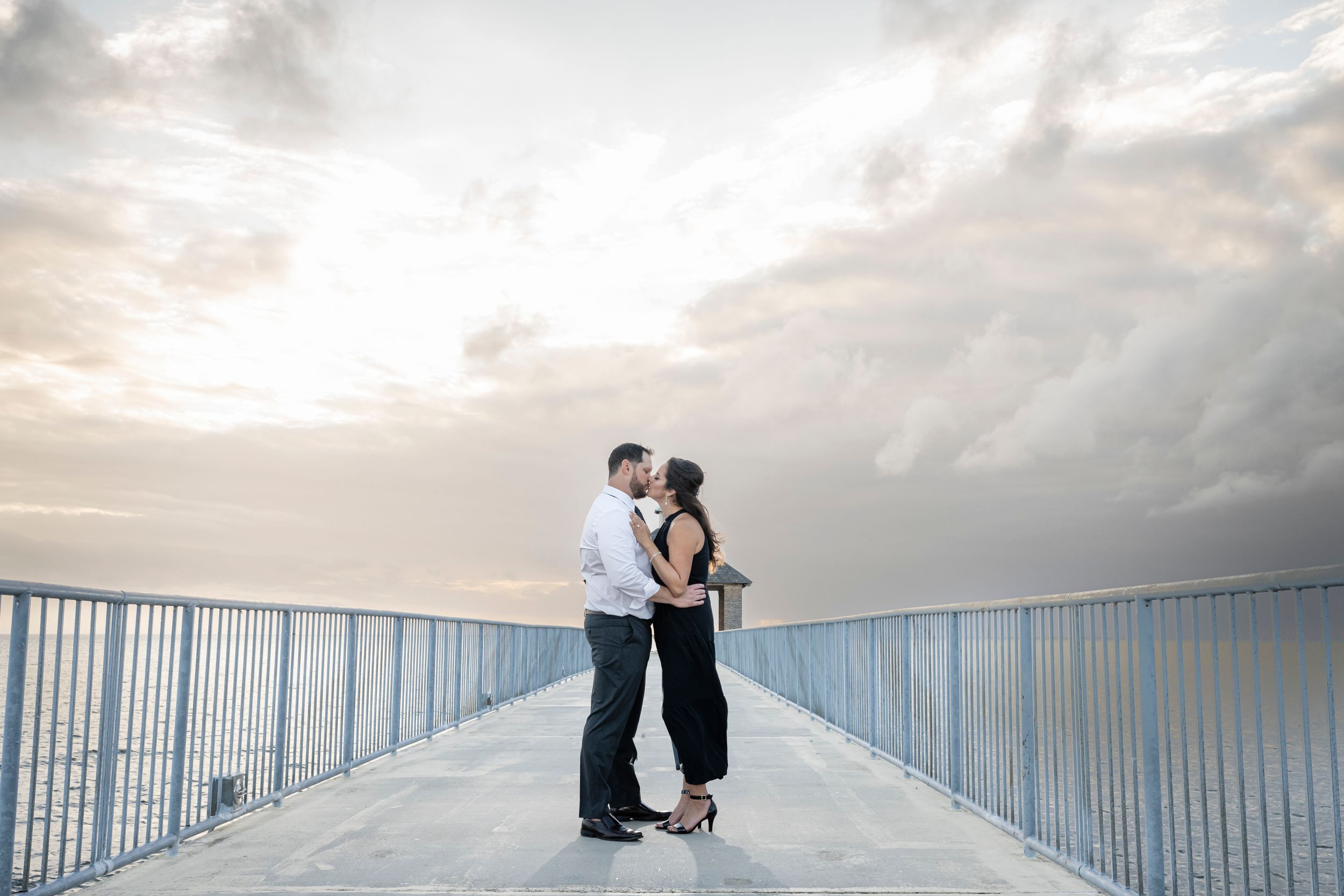 A couple embraces on a pier with a cloudy sunset sky creating a dreamy backdrop.