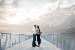 A couple embraces on a pier with a cloudy sunset sky creating a dreamy backdrop.