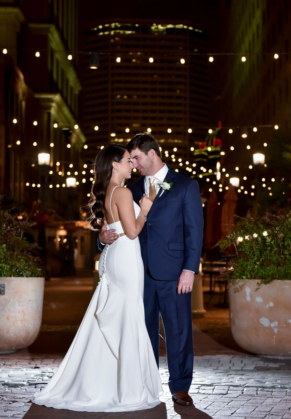 A romantic nighttime wedding photo with string lights and urban architecture in the background.