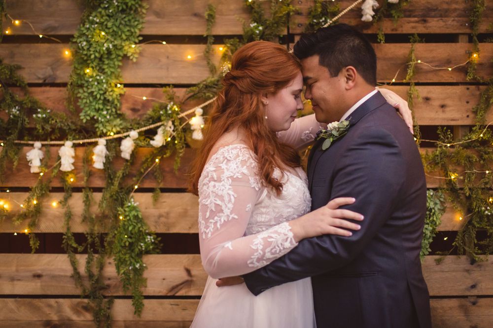 A romantic wedding couple embraces in front of a rustic wood wall with string lights and greenery.