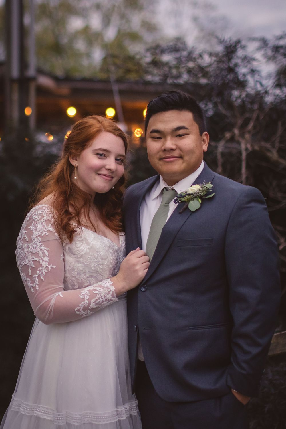 A newlywed couple poses together outdoors with string lights glowing in the background.