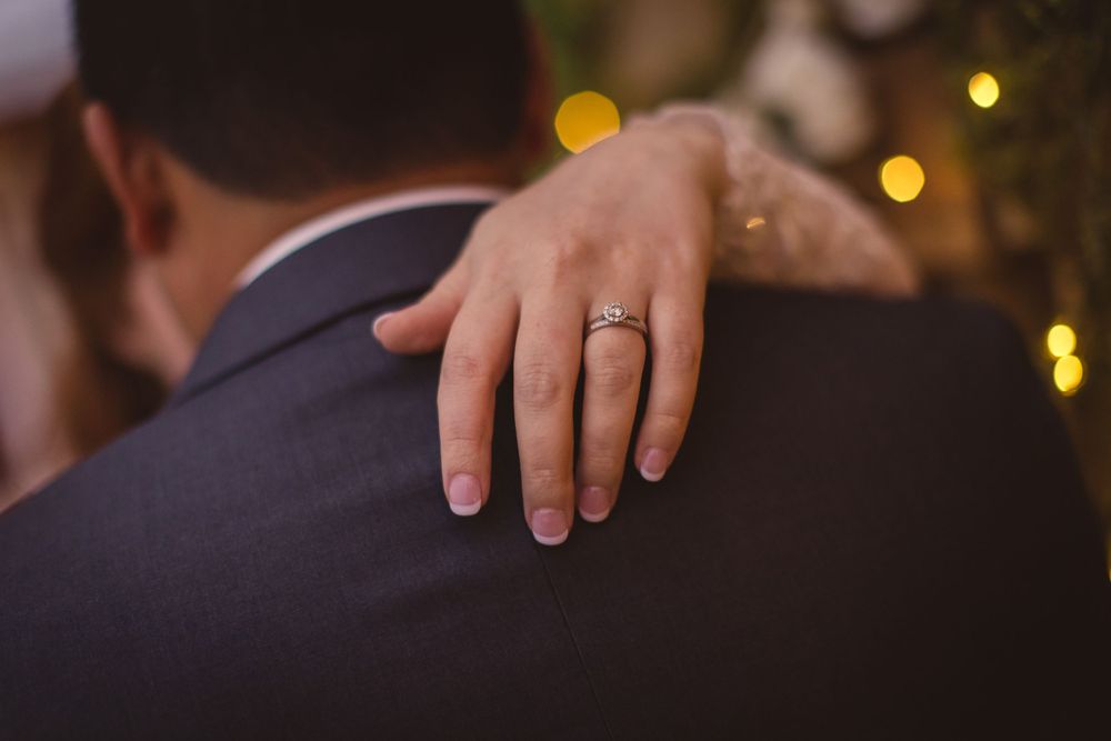 A close-up of a bride's hand with wedding ring resting on groom's navy suit jacket.