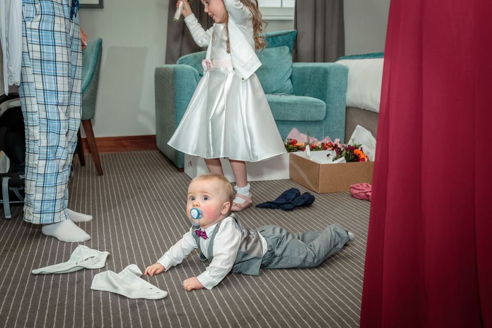 young baby in a shirt and waistcoat lying on his stomach the hotel floor as the bridal prep continue around him