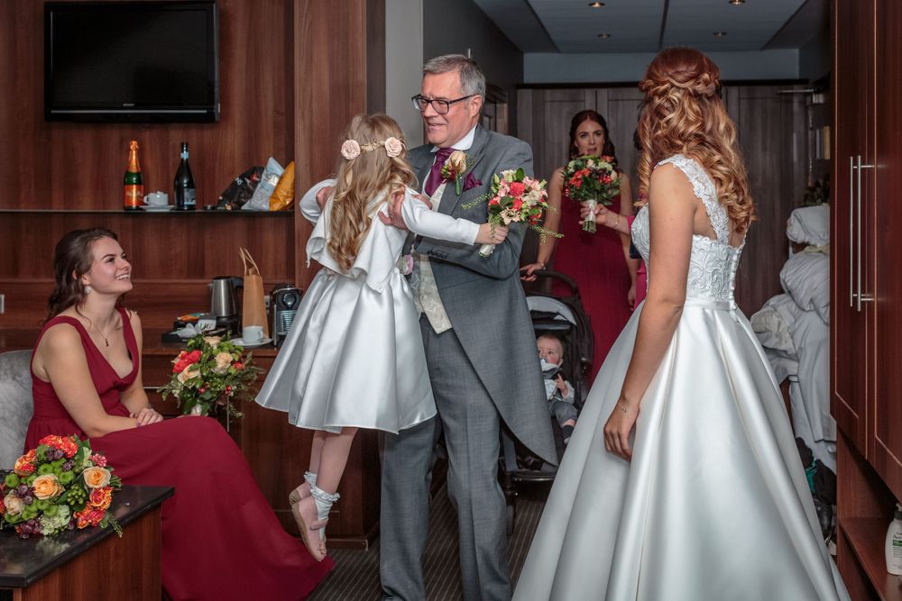 father of the bride lifts up the young flower girl watched by the bride and bridesmaids