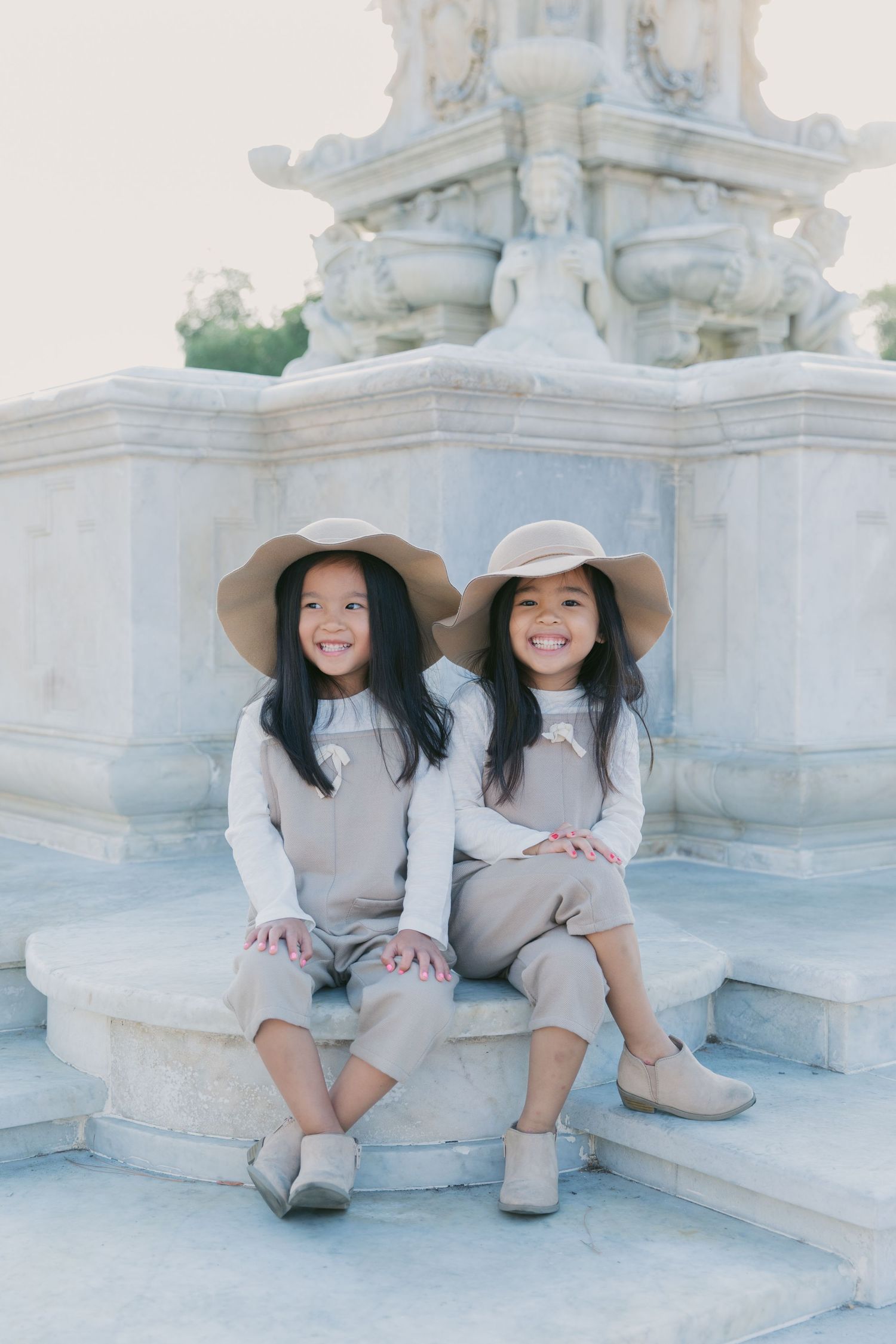 Two people in beige outfits and sun hats posing by a white stone structure.