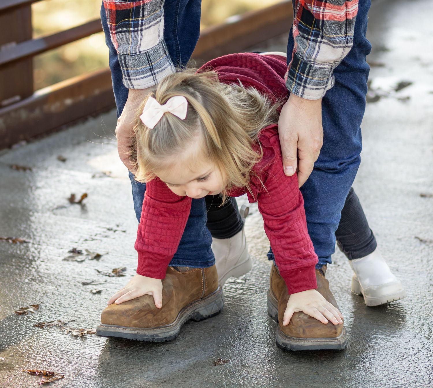 Young blonde girl in red hoodie bending down touching adult boots on wet pavement.