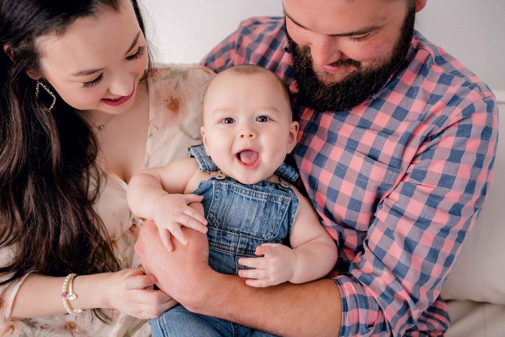 Happy baby in denim overalls smiling while being held by parents on white couch.