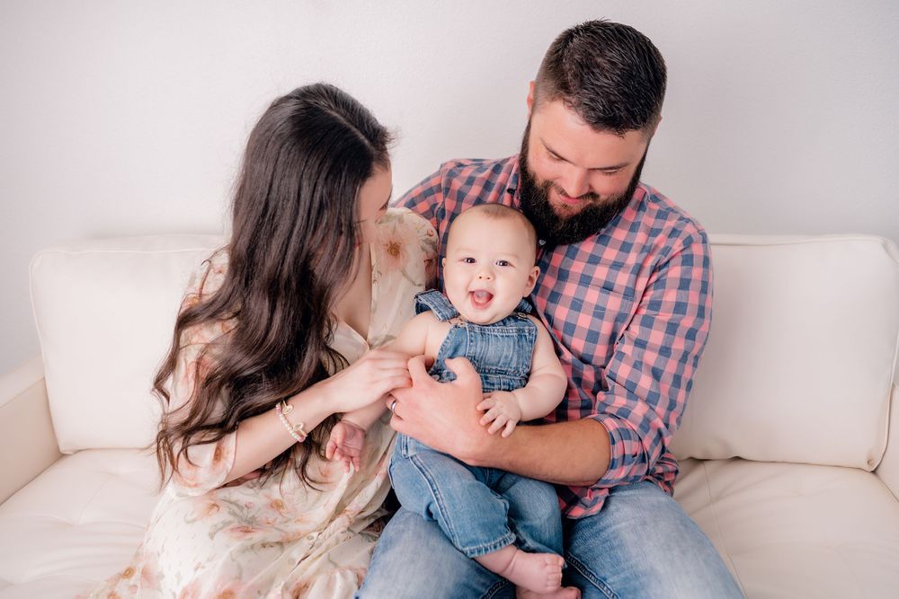 Family sitting together on white couch with baby wearing blue denim overalls.