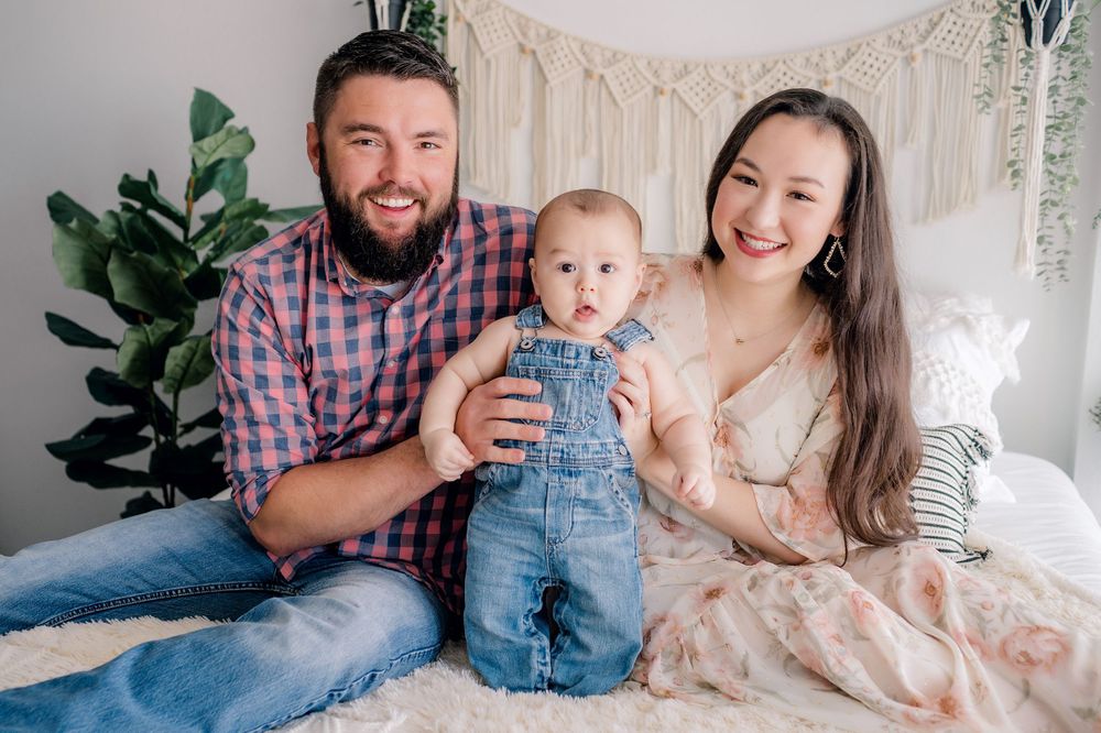 Family portrait on white bed with macrame backdrop and fiddle leaf fig plant.