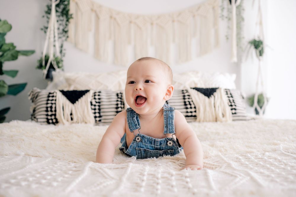 Baby in denim overalls sitting on white bed with macrame wall hanging and plants.