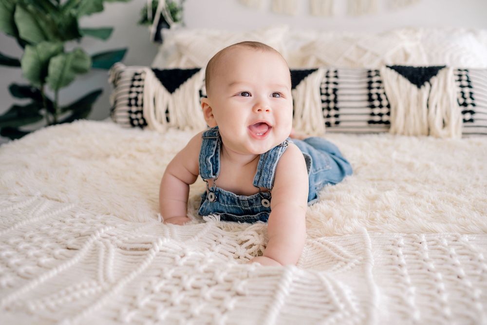 Baby wearing denim overalls lying on white textured bedding with decorative pillows.