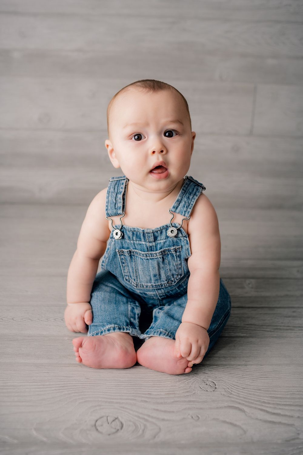 Baby in denim overalls sitting on gray wooden floor during photo session.