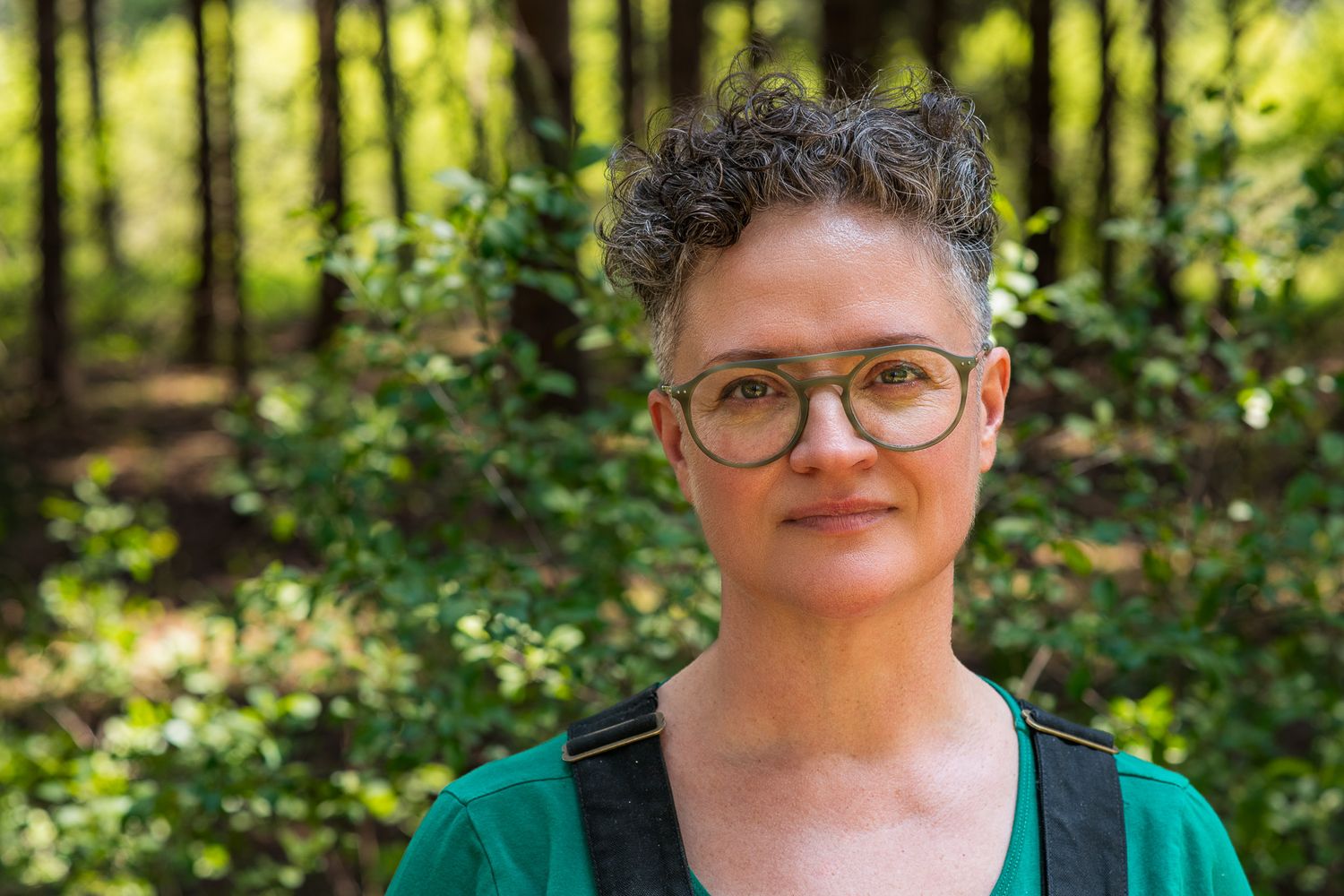 a grey haired woman in grey glasses and overalls stands in the woods