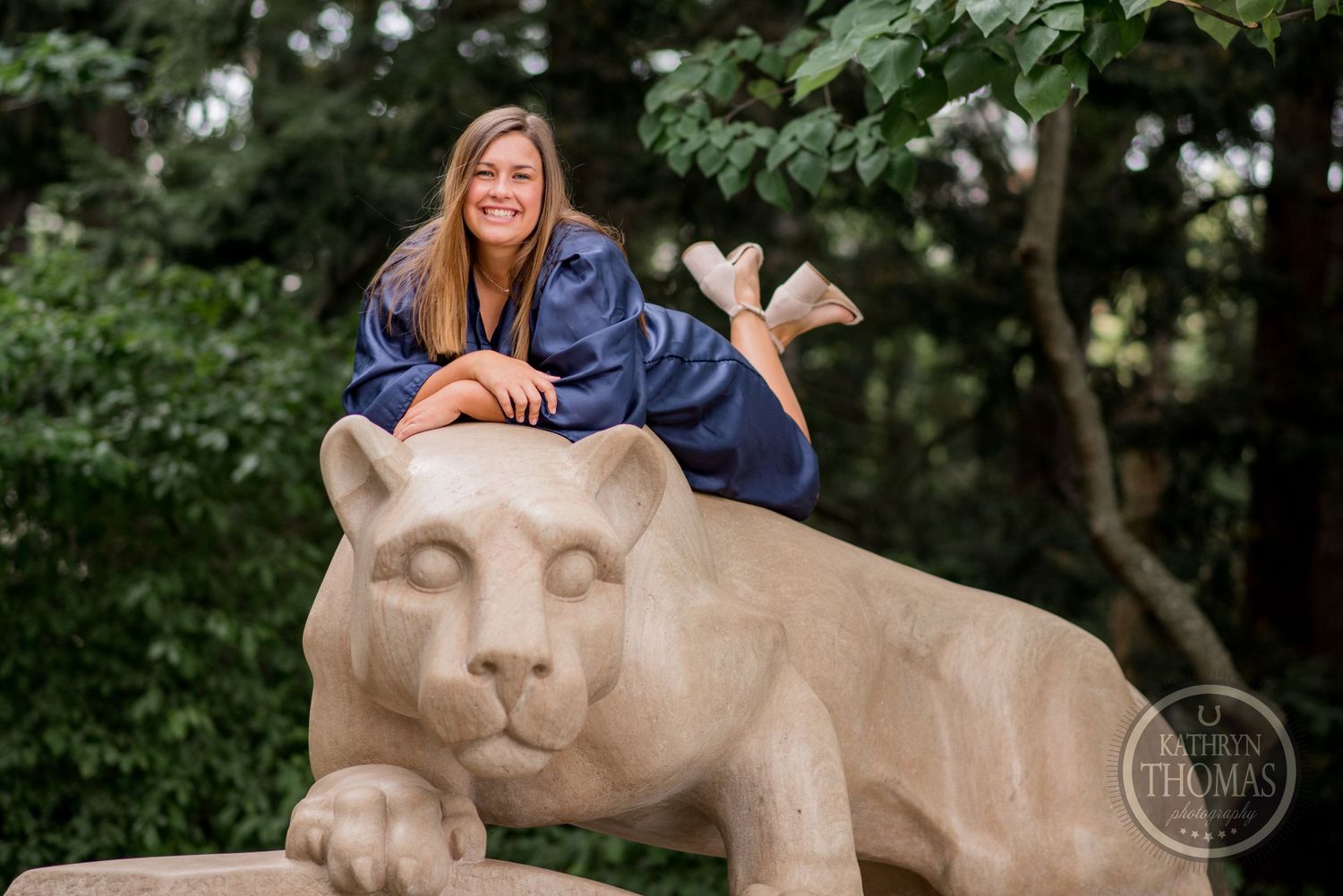 Graduate in blue academic regalia playfully poses on top of Penn State lion statue.