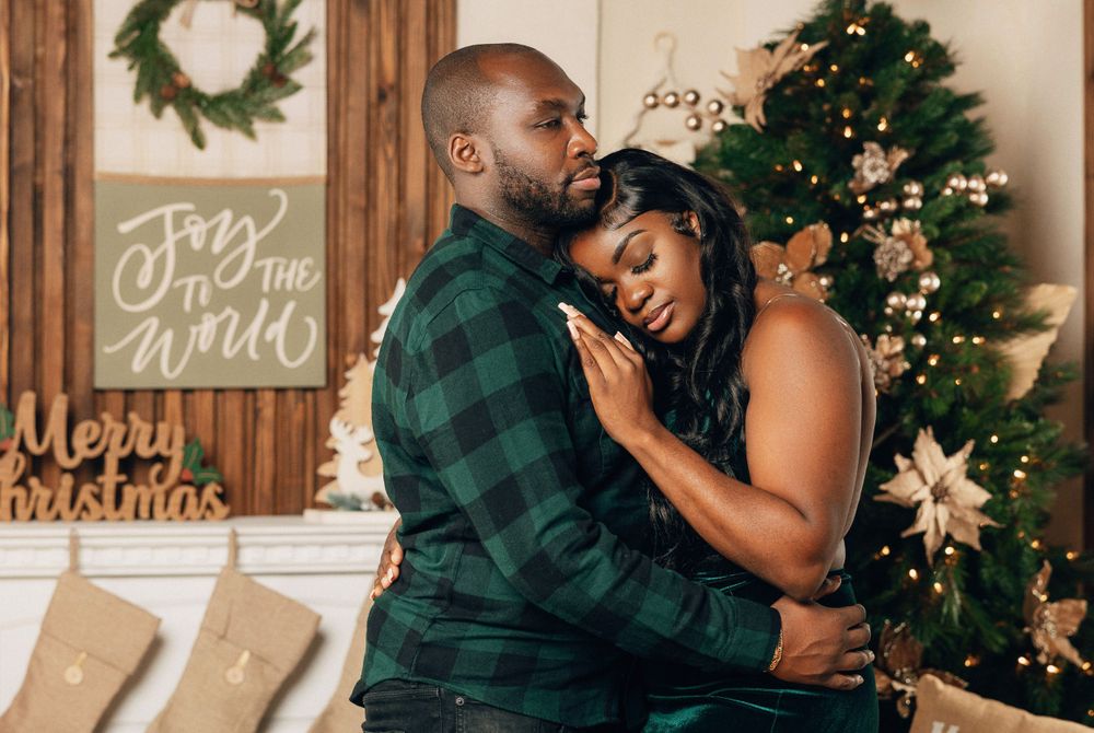 Couple embracing tenderly in front of Christmas tree with holiday decorations and Merry Christmas sign.