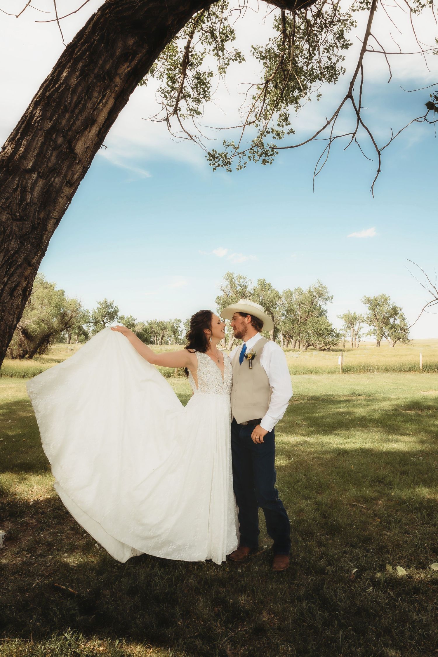 A couple poses under a large tree on their wedding day in a rustic countryside setting, her white dress flowing in the breeze.