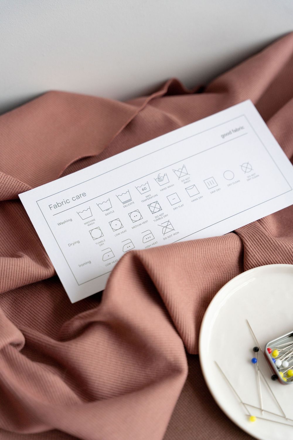 Fabric care instruction sheet displayed on brown textile with sewing pins in ceramic dish.