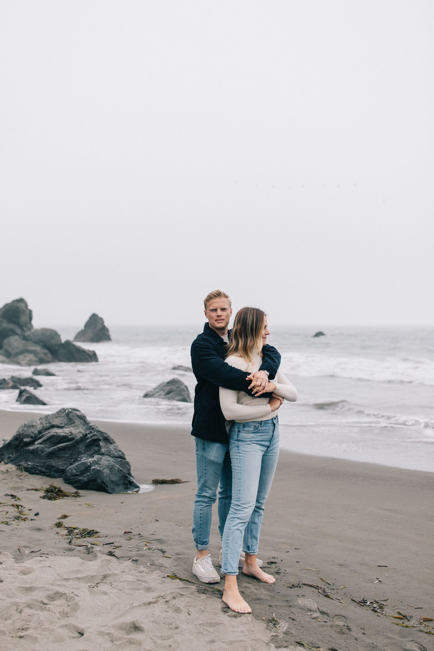 A couple embraces on a moody beach with dark volcanic rocks and ocean waves in the background.
