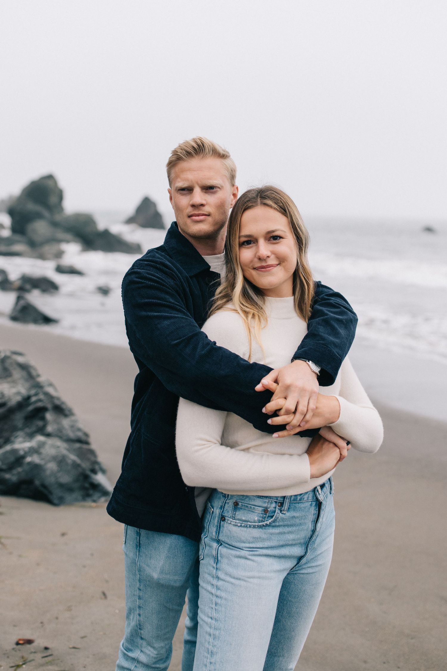 A couple shares an intimate moment on a rocky beach wearing casual denim and neutral colors.