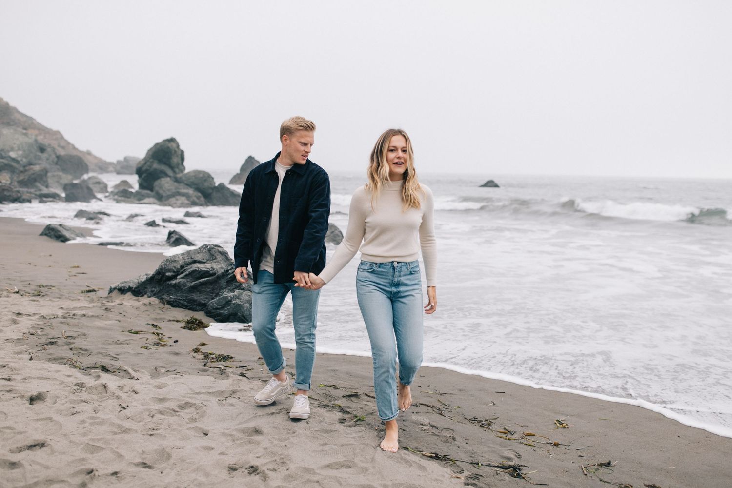 A couple takes a romantic stroll along a misty beach with black rocks and crashing waves.