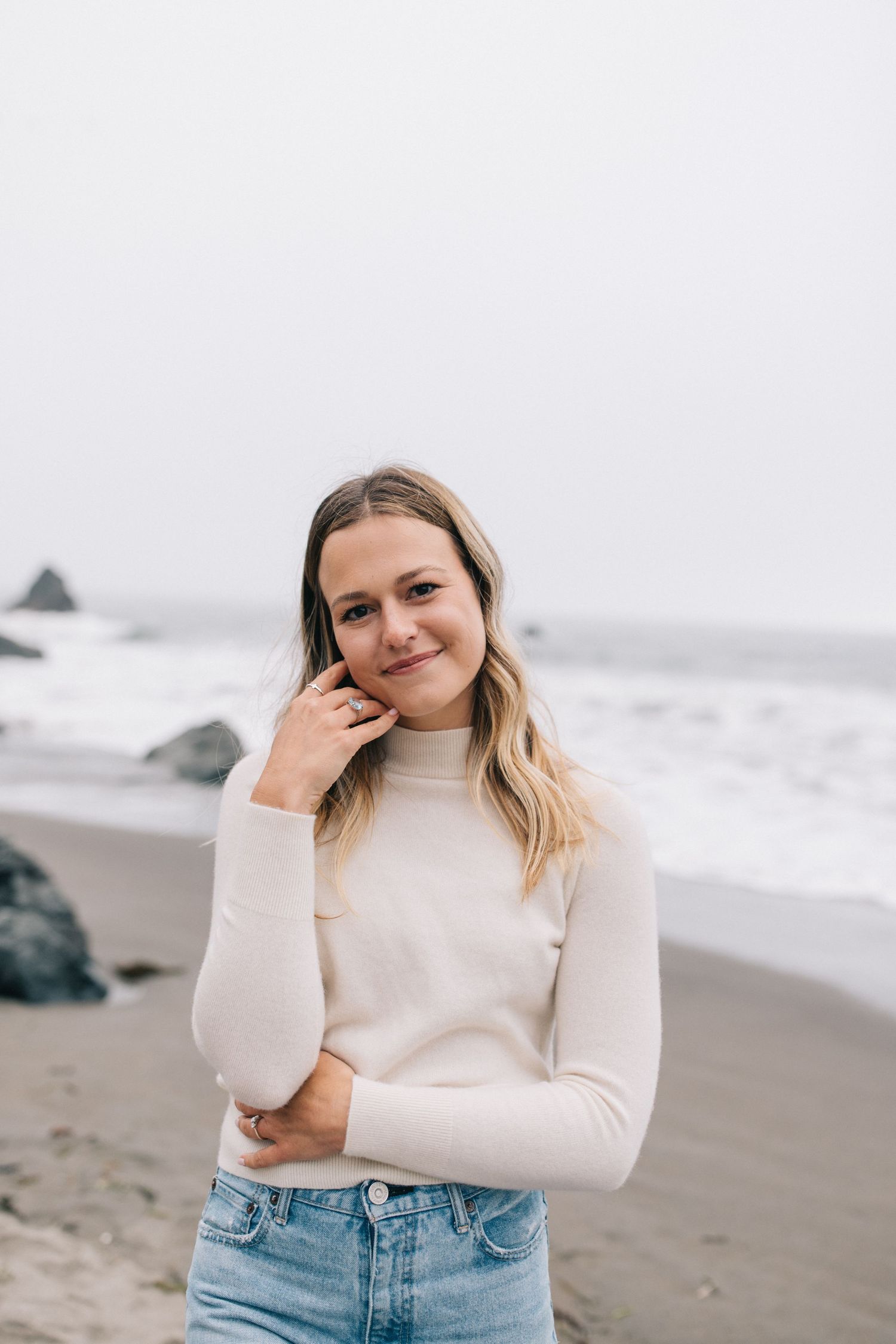 A casual beach portrait showing a woman in a cream sweater against a misty coastal backdrop.
