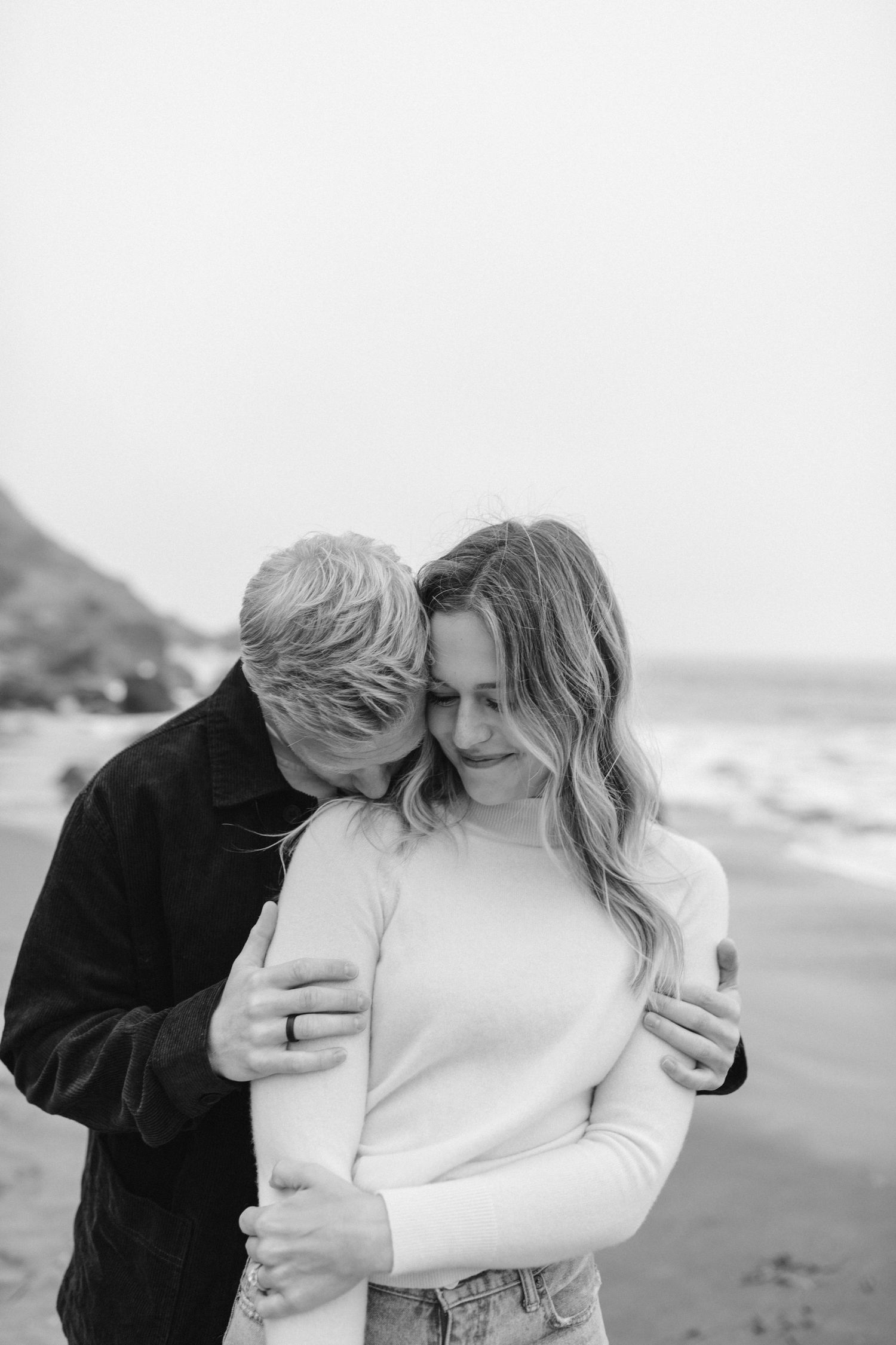 A romantic black and white portrait of a couple embracing on a rocky beach with waves in the background.
