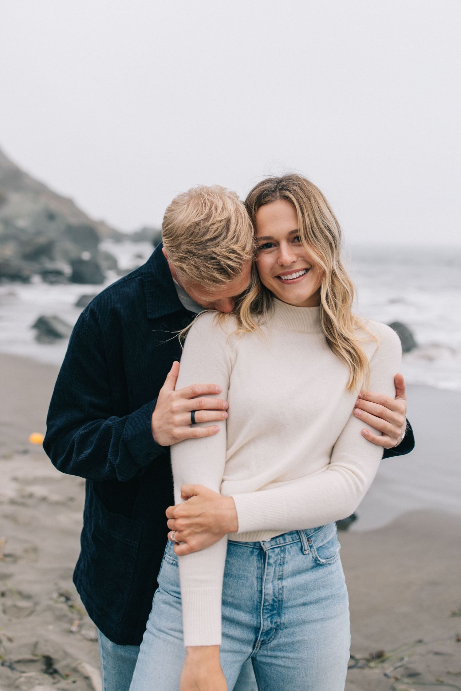 Intimate embrace between couple wearing casual outfits on foggy beach.