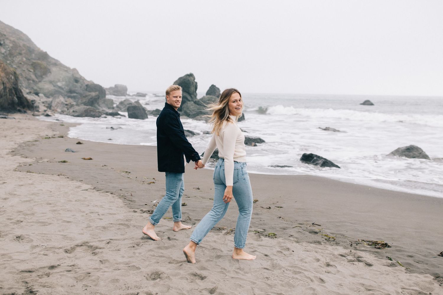 Couple enjoying casual barefoot stroll along rocky coastline on overcast day.
