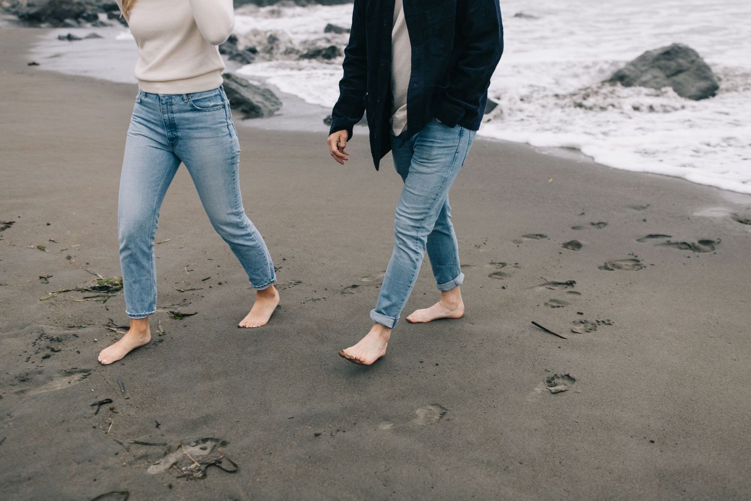 Close-up of barefoot legs walking on dark sand beach near ocean.