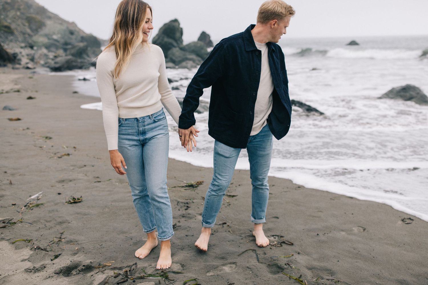 Barefoot couple holding hands while walking along misty coastal shoreline.