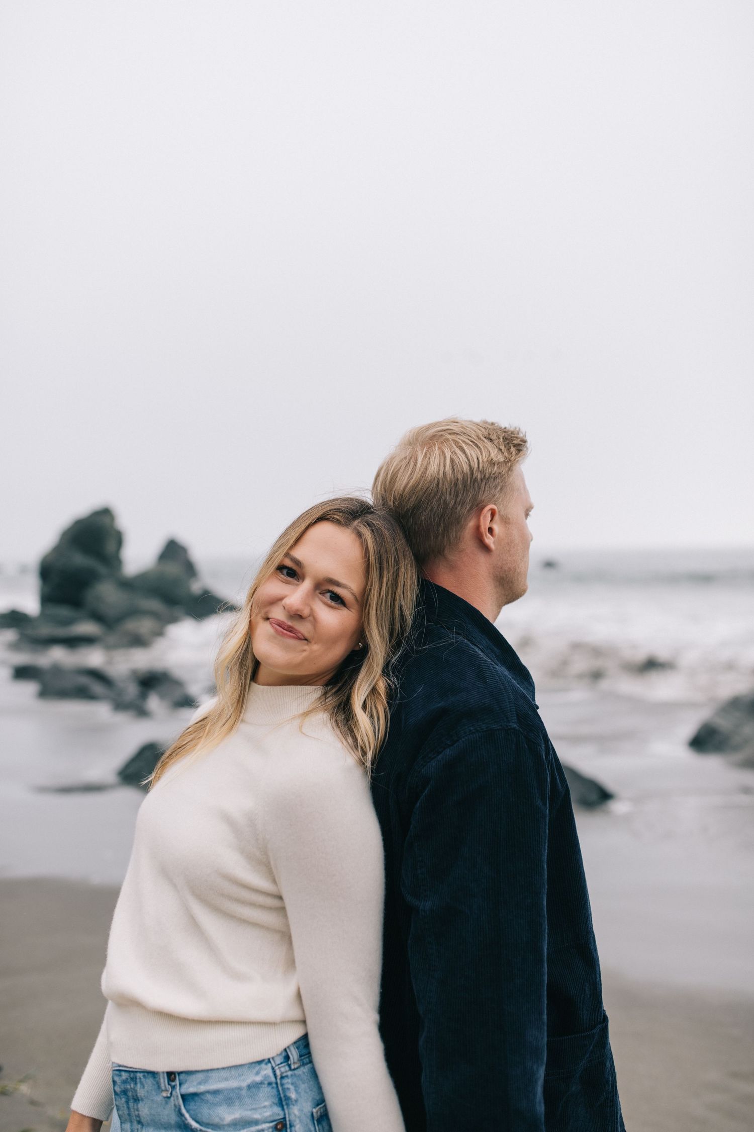 Couple in casual winter outfits posing together on rocky beach.