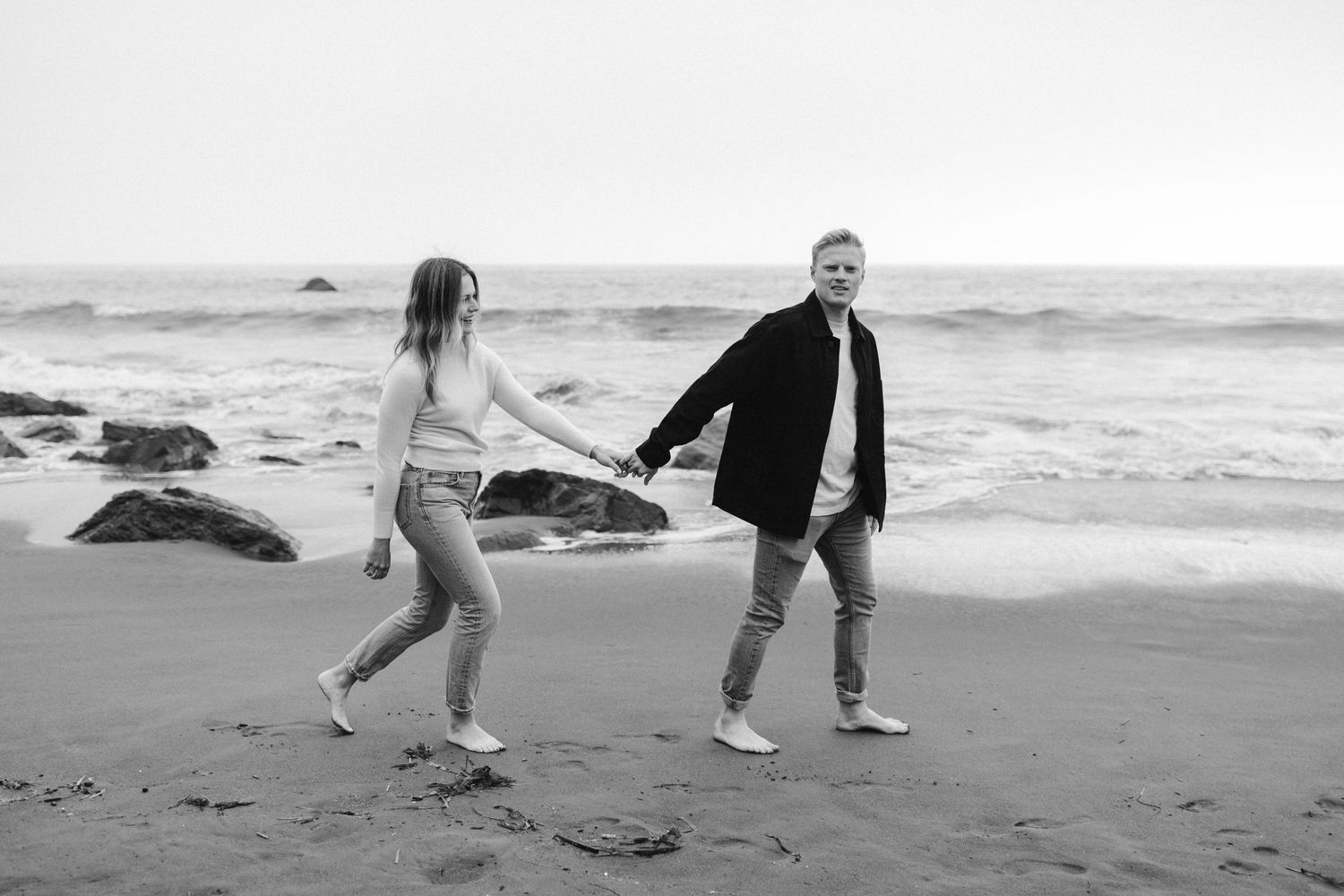 Black and white photo of couple playfully walking on beach near ocean waves.