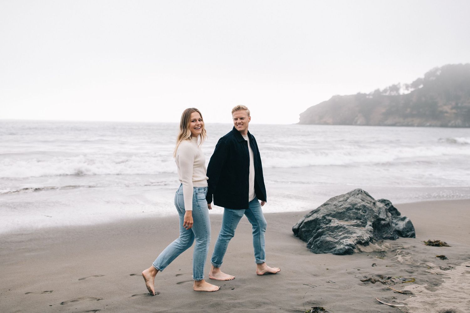 Couple walking barefoot together on a misty beach with rocks and waves in the background.