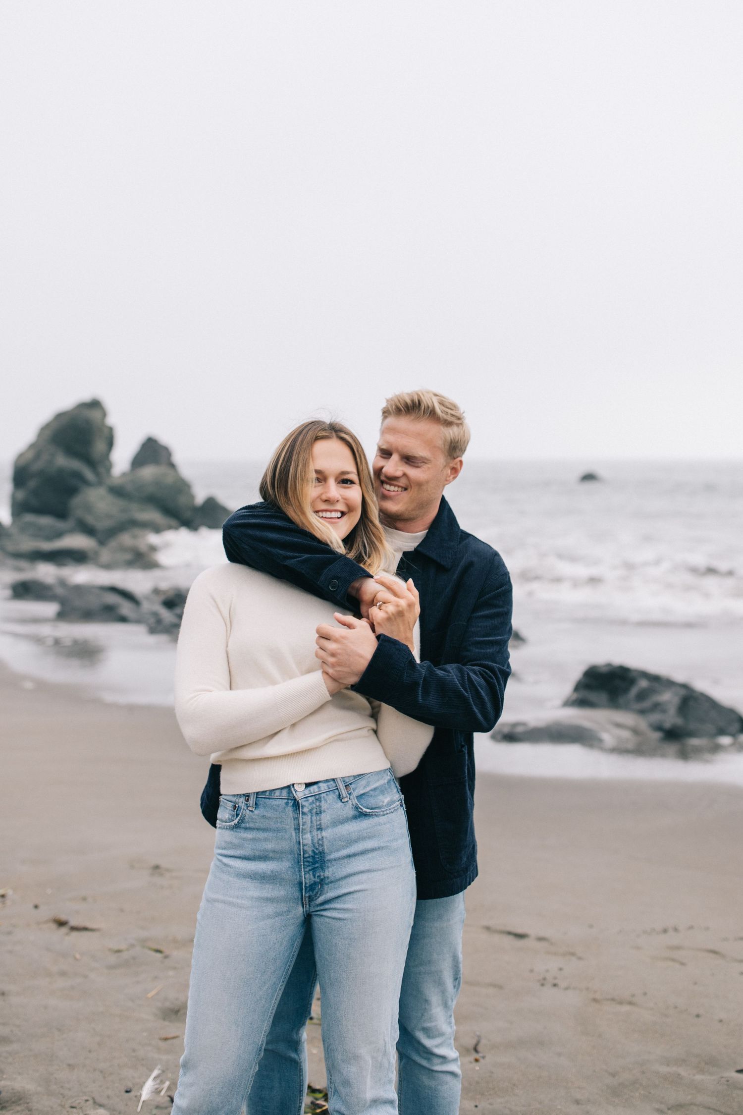 Sweet embrace captured during beach photoshoot with moody coastal scenery in background.