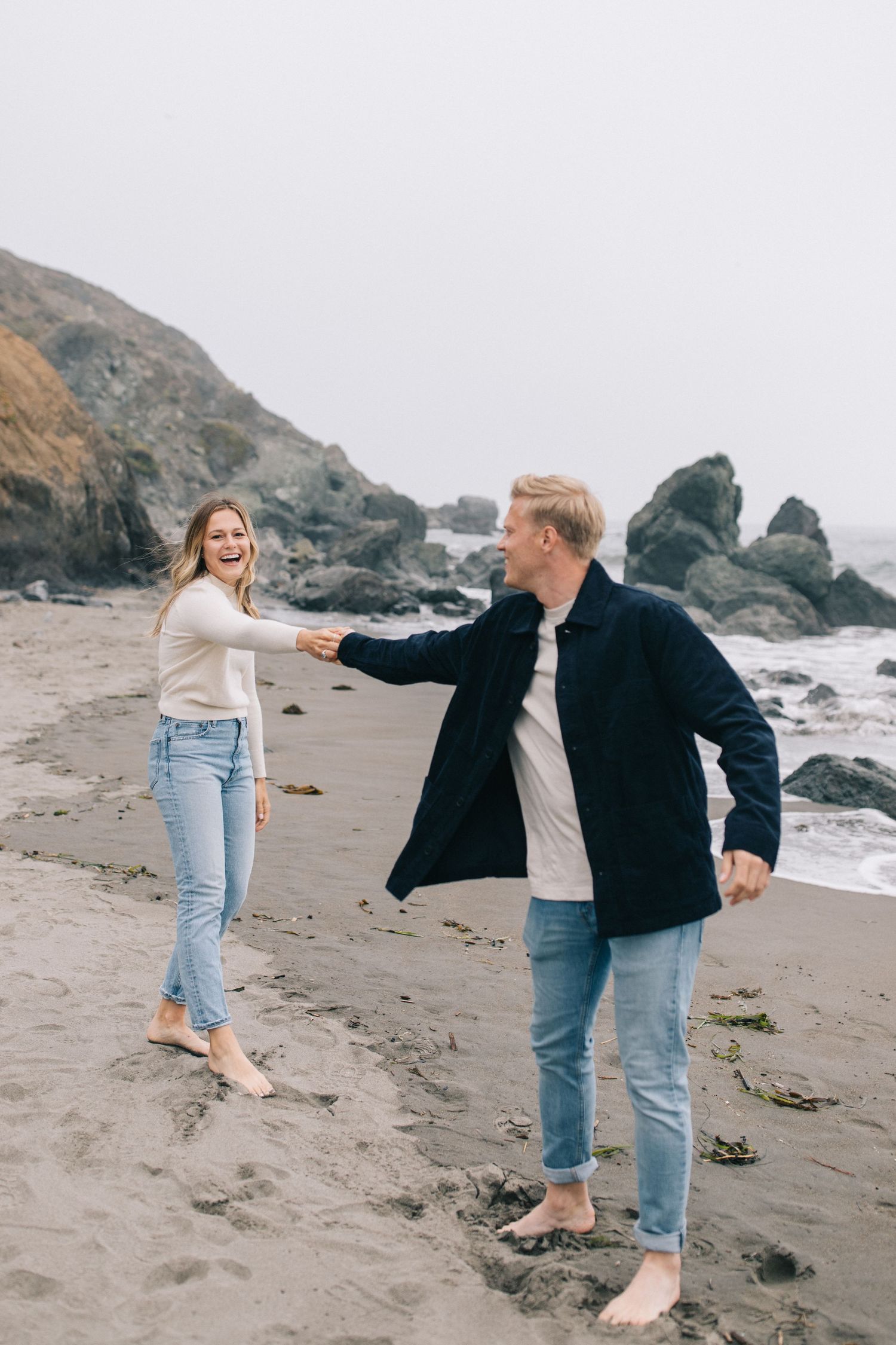 Couple dancing barefoot on beach with coastal rocks and grey skies creating moody backdrop.