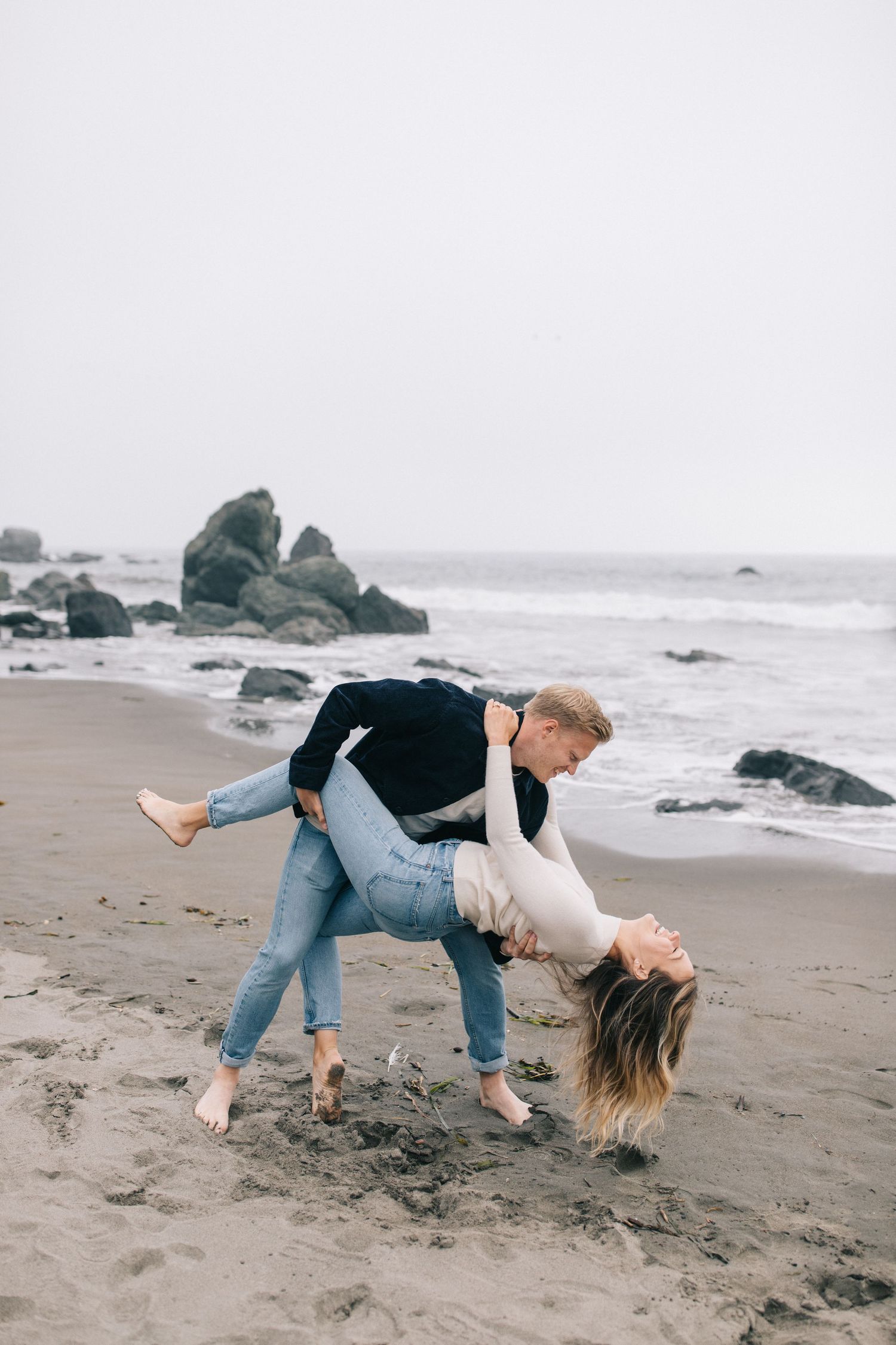 Dramatic dip pose during beach photoshoot with crashing waves and rocky coastline.