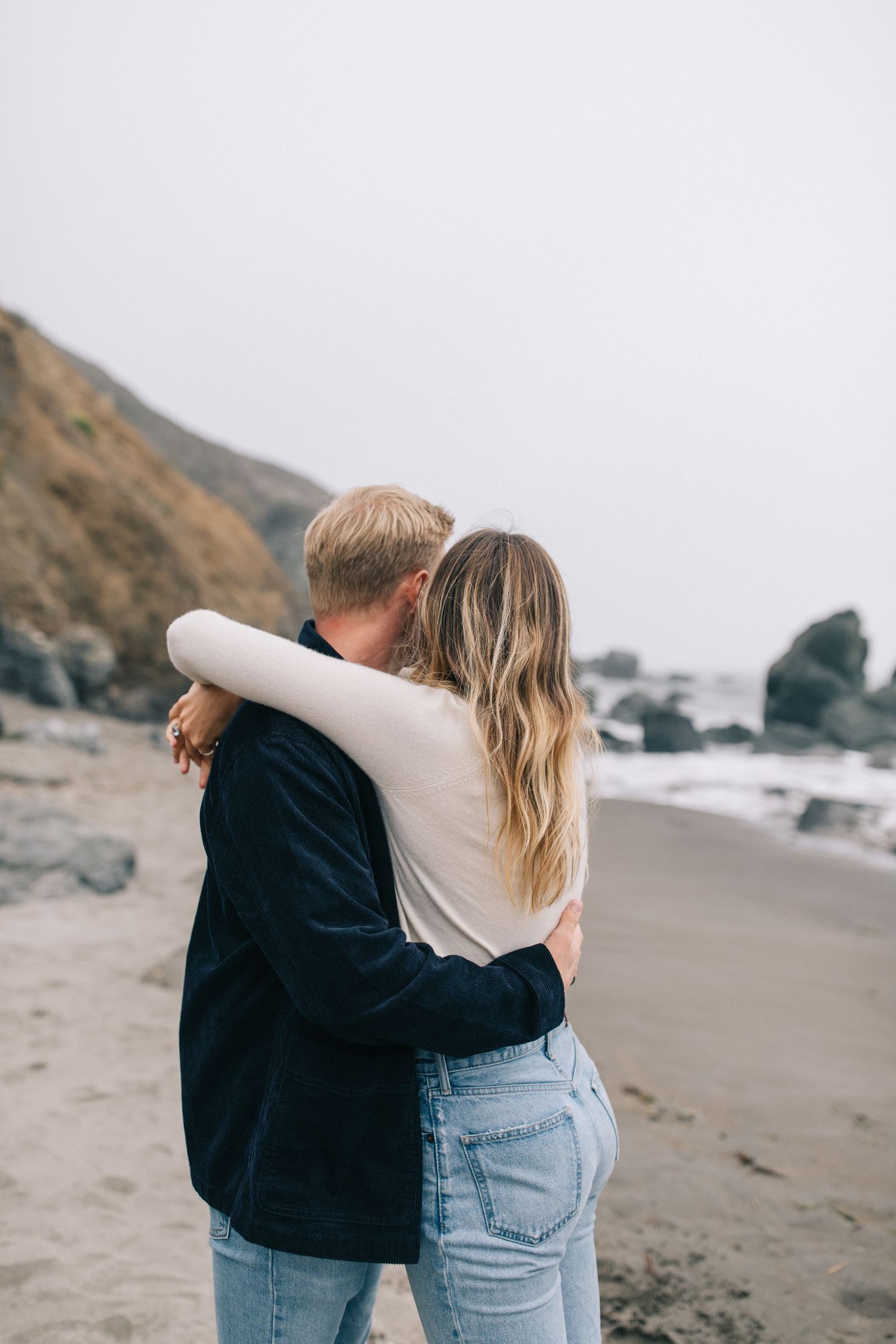 Couple sharing tender moment on foggy beach with dramatic coastline in background.