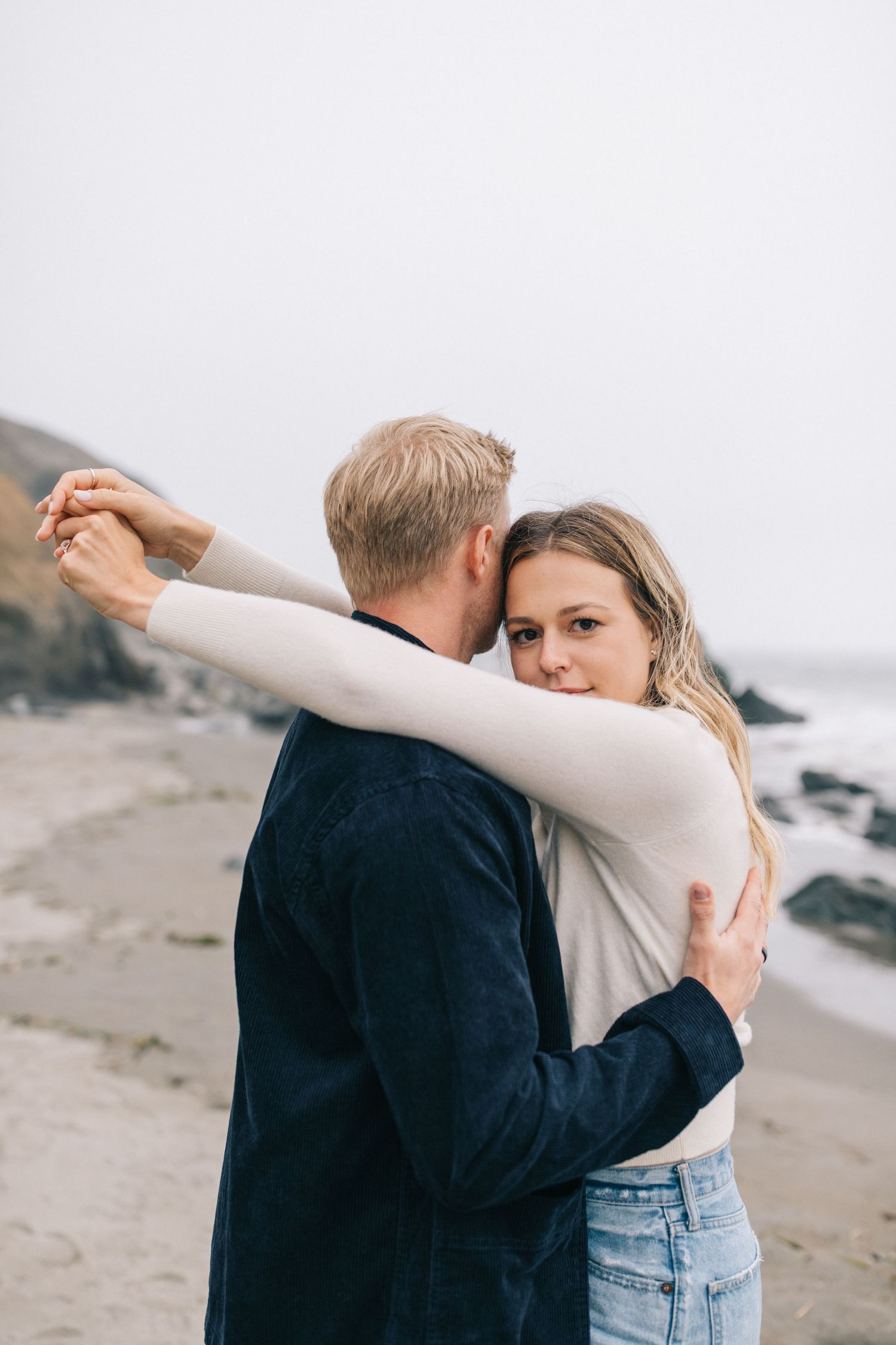 Romantic embrace on a misty beach with coastal cliffs and ocean waves in the background.