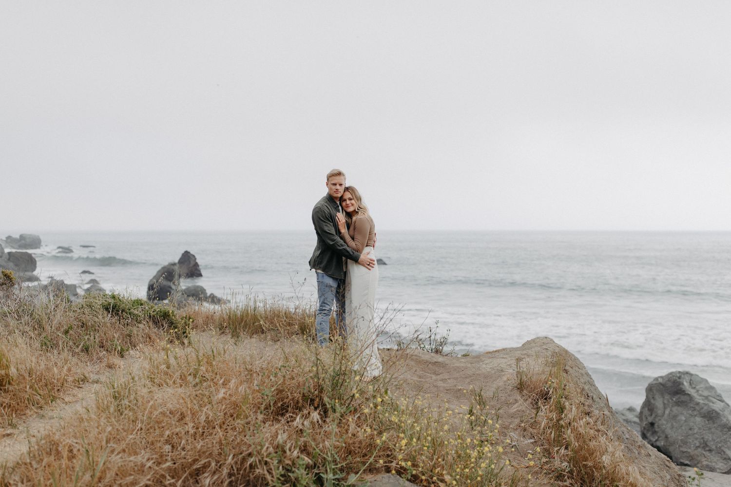 Couple stands together on rocky coastal cliff with crashing waves and moody grey skies in background.