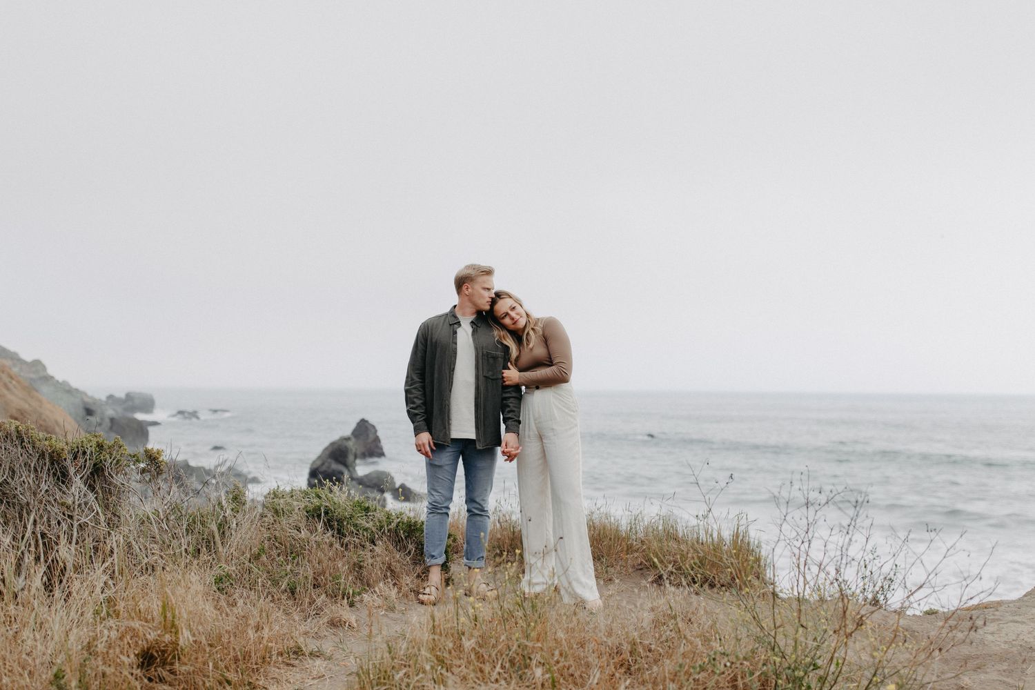 A romantic coastal photoshoot with a couple embracing on a cliff overlooking the Pacific Ocean on an overcast day.