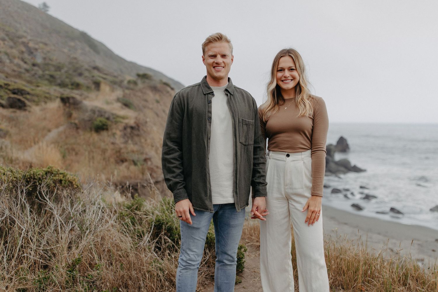 A casual portrait of a couple standing on a coastal path with ocean views.